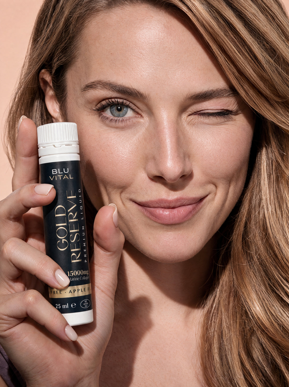 Knipogende vrouw met een BLUVITAL The Gold Reserve-collageenshot van 25 ml naast haar gezicht.